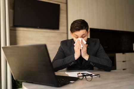 Sick young man at work got allergic to flu, sneezing and fasting, wiping a runny nose with a paper napkin, allergic guy caught a cold at work, respiratory illness sitting in home office, sick leaveの写真素材