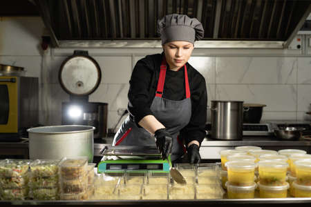 Food delivery in the restaurant. female chef in uniform prepares food in a restaurant and prepares it in disposable dishes. Places portions of rice in a disposable dish on a scale.の写真素材