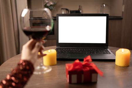 A blank laptop screen stands on the table. A womans hand raises a glass of red wine. concept of dating online at a distance.の写真素材
