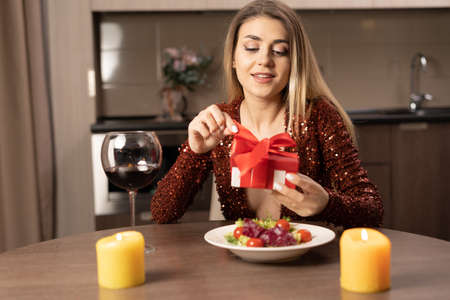 smiling young woman opening a valentine gift sitting at the table in a cozy kitchen. Giving gifts to your loved ones concept. Webcam portrait of a girl, wife, housewife for an online video call.の写真素材
