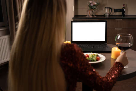 blank computer screen over a womans shoulder, a woman is having dinner at home in a beautiful dress and holding a glass of red wine in her hands. video call concept.の写真素材