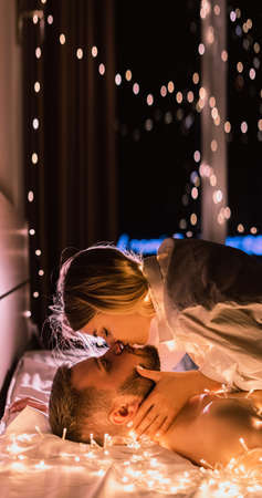 Close up portrait woman and man at night lie at home on the bed in the light of garlands. authentic atmosphere of Valentines Day or Christmas. lady kissing boyfriend.Baner. copy space.の写真素材