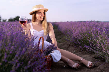 A young beautiful girl sitting among the lavender bushes holds a glass with red wine in her hands in the summer at sunset. Wine tastingの写真素材