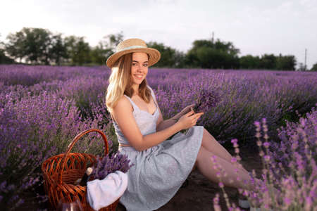 A young beautiful girl sitting among the lavender bushes holding a bouquet of flowers in the summer at sunset. field of blooming lavender and a lady in a hat.の写真素材