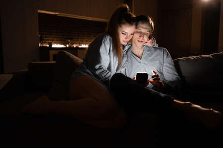 Happy young woman and man use a smartphone together, sit at home on a cozy sofa, at night wife and husband look at the phone screen, online shoppingの写真素材