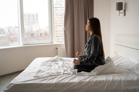Portrait of a long-haired brunette in pajamas, meditating, sitting on a soft bed in the morning at home or in a hotel, large windows overlooking the city. copyspace.の写真素材