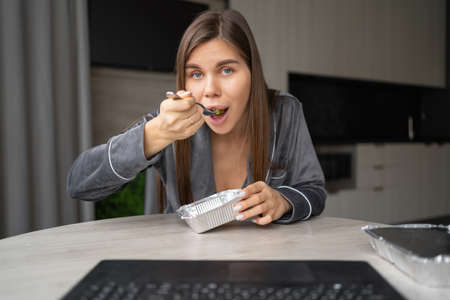 Happy woman enjoying a delicious takeaway lunch at home. A beautiful girl sits at a table and eats from a food container. Food delivery service concept. makes a video call on a laptop. webcam viewの写真素材