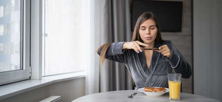 happy young woman sitting at home at the table having breakfast pancakes with orange juice. Takes a photo of your food with your smartphone. copy spaceの写真素材