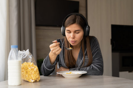 Young woman in pajamas having breakfast with corn flakes with milk at home. Listen to music with...
