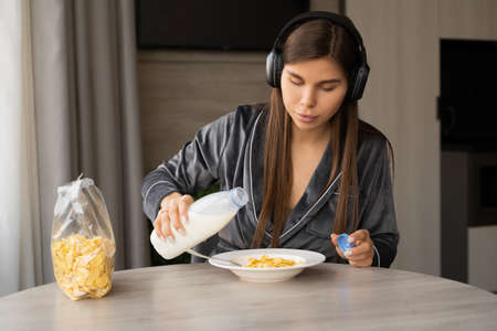 A happy woman pours milk into a bowl of cereal during breakfast and listens to music on wireless headphones. copyspace.の写真素材