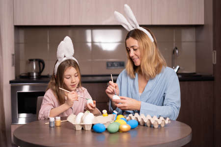 Mother and daughter paint Easter eggs at home in the kitchen. Happy family is preparing for Easter. Cute little girl with bunny ears on Easter day. Preparation for the holiday. copyspace.の写真素材