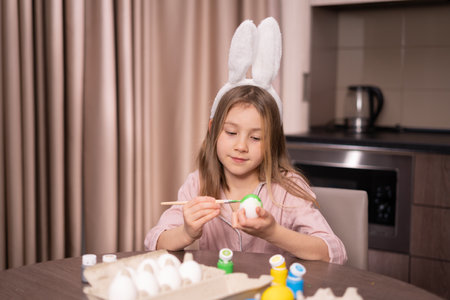 A little girl with rabbit ears paints eggs for Easter at home in the kitchen.の写真素材