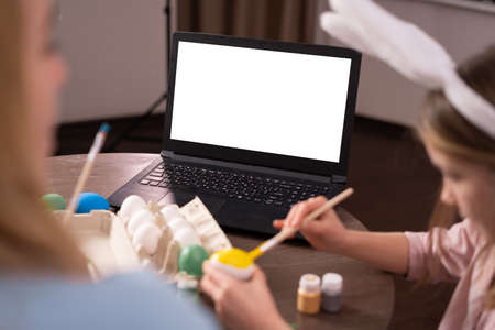 A laptop with a blank screen on a table festively decorated for Easter. close-up of hands paint chicken eggs for the holiday.の写真素材