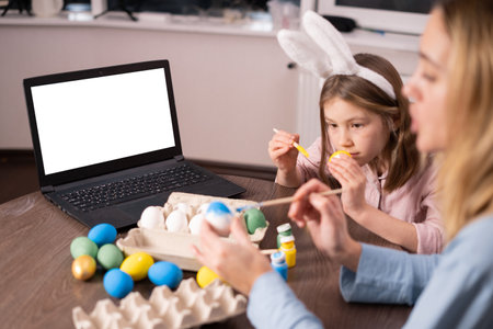 laptop with a blank screen on table festively decorated for the Easter holiday.Mother and daughter paint Easter eggs at home in the kitchen. Happy family preparing for Easter. little girl bunny earsの写真素材