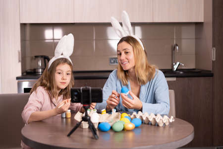 mother and daughter in a bandage on the ears of a rabbit are sitting in the kitchen at home. They decorate Easter eggs with paints and take selfies on their smartphones, smiling cheerfully.の写真素材