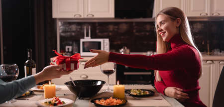 Couple in love having fun celebrating valentines day enjoying a romantic dinner, happy young woman receiving a gift box from a loving manの写真素材