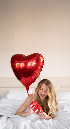 Valentine's Day, Women's Day. A young caucasian brunette lies in bed and celebrates valentine's day, holding a heart-shaped balloon and a red box in her hands. copyspace.の写真素材