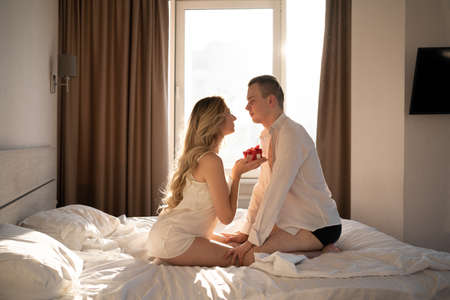 Romantic morning at the hotel. A man and a woman on the white sheets of a warm hotel room in bed. A woman gives a man a gift in a red box. valentine's day, anniversary or anniversary.の写真素材