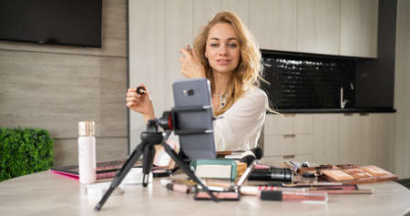 A beautiful young woman blogs about makeup and beauty products. Gorgeous lady removes makeup tutorial with makeup lipstick. Smiling girl teaches makeup using her phone.の写真素材
