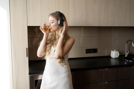 A young woman in casual clothes is relaxing with music in headphones in a cozy home, enjoying the morning sun from the window and hot tea.の写真素材