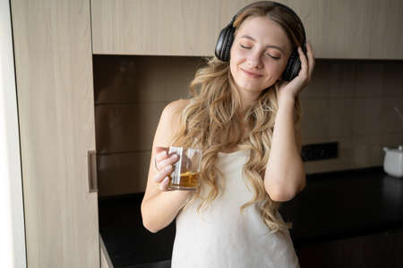Caucasian woman in a white shirt with headphones on her head dances in the kitchen at home in the morning. A woman listens to music while drinking her morning tea.の写真素材