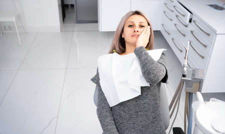 A young caucasian girl in casual clothes touches her mouth with her hand with a painful expression on her face due to a toothache or dental disease of the teeth sits on a chair at the dentist. copy spaceの写真素材