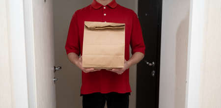 Courier delivery. deliveryman in red uniform with a package at the door, close-up. courier with restaurant food. man with takeaway food package.の写真素材