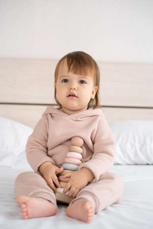 Eco-friendly toys for kids. a little girl sits at home on the bed and holds a modern wooden colorful pyramid with rings in her hands. development of logical thinking in children. Place for textの写真素材