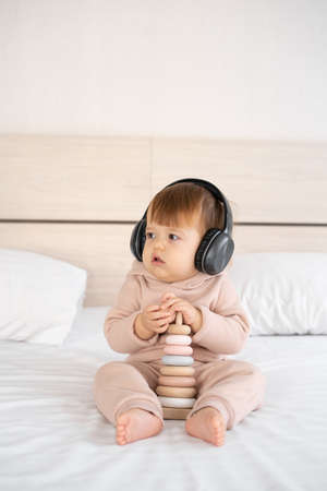 cute child girl preschool listening to music in headphones, playing wooden pyramid toy, sound for children, happy childhood concept, child on white bed in headphones.の写真素材