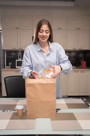 Smiling cheerful woman unpacking home delivery fast food tasting while standing in the kitchen ordering lunch or dinner.の写真素材