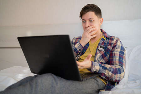 male remote worker with a laptop sits at home during the day on the bed in casual clothes, covers his mouth with his hand, shocked and afraid of a mistake. surprised expression. copyspace.の写真素材