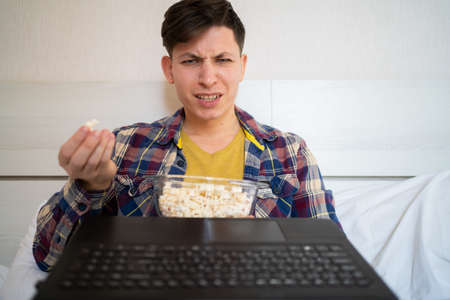Photo of tense angry guy holding popcorn with open mouth while sitting on bed at home. online cinema concept. webcam viewの写真素材
