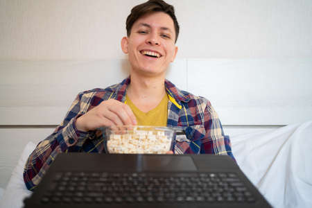A young guy is relaxing at home and watching a movie on his laptop. eats popcorn and laughs merrily. online cinema concept. webcam viewの写真素材