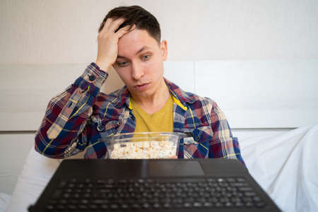 young caucasian man sits on the bed at home and watches a movie in an online cinema. Holds popcorn in hands, emotion of disappointment and surprise. webcam view. copyspace.の写真素材