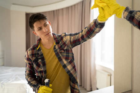 A young hipster guy washes a mirror at home. caucasian man in gloves wipes mirror with detergent sprayer and cloth, does housework. Men's washing mirror in the living roomの写真素材