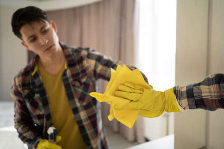 A young hipster guy washes a mirror at home. caucasian man in gloves wipes mirror with detergent sprayer and cloth, does housework. Men's washing mirror in the living roomの写真素材