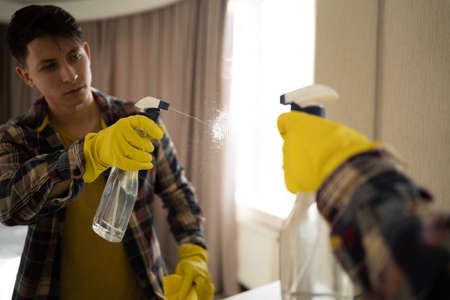 A young man washes a mirror in a home hotel wearing yellow rubber gloves. copy spaceの写真素材