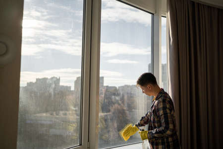 caucasian man in yellow rubber gloves washes windows at home during the day in casual clothes. copyspace.の写真素材