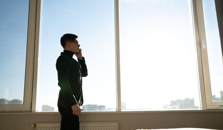 a male businessman stands in the office near the panoramic window making a call using his mobile phone. Digital technology applications and solutions for corporate business development.の写真素材