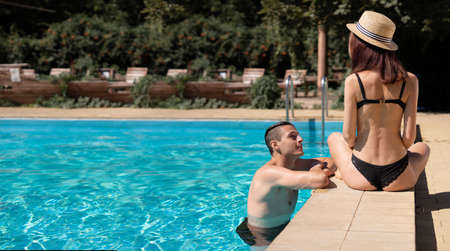 Young adult woman sitting on the edge of the pool laughing and flirting with a man floating in the water. The concept of romance and flirting while on vacation at the resort.の写真素材