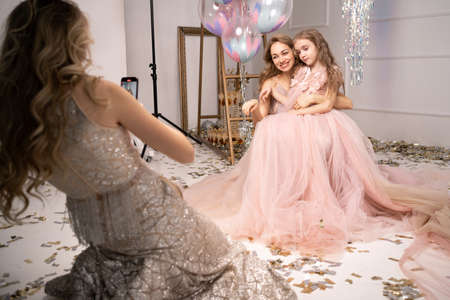 photo session on a smartphone at a professional photo studio. a woman and a child in beautiful long dresses pose for a mobile photographer. photo session of mother and daughter.の写真素材