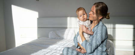 A young woman sits on a bed and holds a baby. good morning. The concept of maternity leave and motherhood. defocusの写真素材