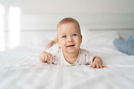 Cute little caucasian baby lies in bed. baby care concept. light background. small child. cheerful emotions. 4 months old boy lying on his stomach in bed with white lines. looking at the cameraの写真素材