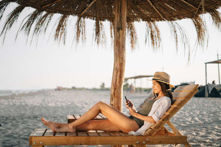 Vacation by sea, weekend on t beach, a beautiful tanned lady in a hat looks at the smartphone screen, sits on a sun lounger on the beach under a straw umbrella. vacation concept. copyspace.の写真素材