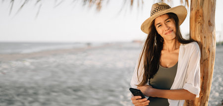 Vacation at sea, weekend on the beach, a beautiful tanned girl in a hat in the camera holds a smartphone in her hands, stands on the beach under a straw umbrella. close-up portrait. Banner. copy spaceの写真素材