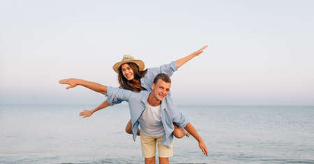 Happy man giving piggyback ride to his woman and laughing at tropical beach. Smiling guy in love carrying on back her girlfriend and having fun. Joyful couple enjoying summer at sea with copy space.の写真素材