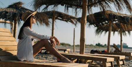 young caucasian woman in a swimsuit resting in the summer on a sandy beach sits in sunglasses on a sun lounger under a straw umbrella. copyspace.の写真素材