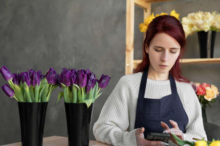 A smiling florist in a blue apron checks orders on the site using apps and a mobile phone while sitting at a table with fresh flowers. The concept of a small flower delivery business. copyspace.の写真素材