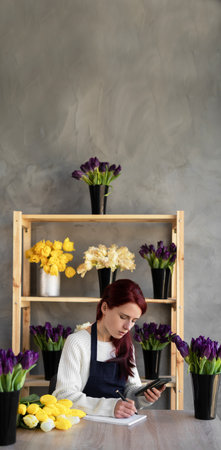 A smiling florist in a blue apron takes orders online using apps and a mobile phone while sitting at a table with fresh flowers. Small business concept selling flowers. banner. copyspace.の写真素材