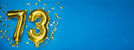 golden yellow foil balloon on blue concrete background number seventy three. Birthday or anniversary card with 73 inscription. Anniversary celebration. Banner.の写真素材